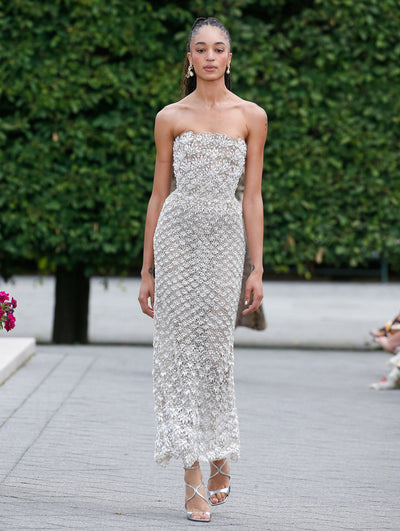 A model is wearing a strapless, fitted gown with an intricate pearl design, featuring delicate floral patterns throughout. The dress has a scalloped neckline and a midi-length hem.