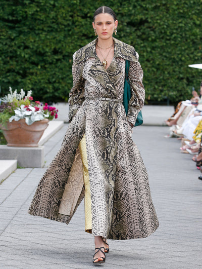 A model is wearing a long, belted snakeskin-print coat dress with a structured collar and wide, flowing skirt.