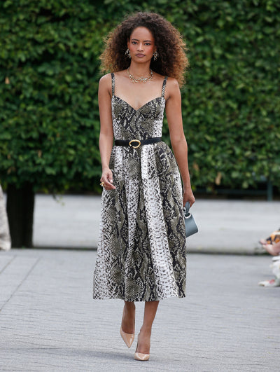 A model is wearing a midi dress with a snakeskin print, featuring thin straps, a fitted bodice, and a flared skirt. The dress is cinched at the waist with a black belt that has a gold buckle.