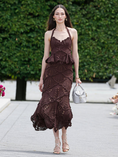 A model is wearing a halter-neck, chocolate brown crochet top with a fitted bodice and a matching chocolate brown crochet scalloped hem skirt. 