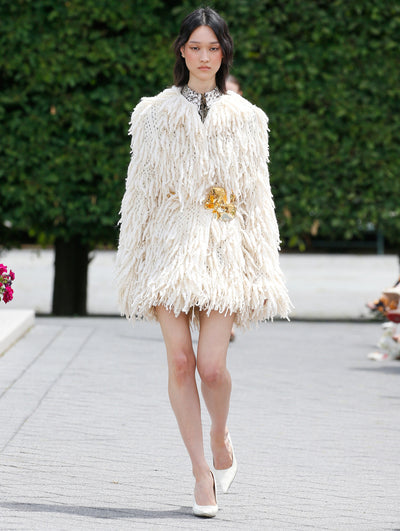 A model is wearing a cream-colored, textured coat with a high neckline and a fringed, feathery design that creates a voluminous silhouette. 