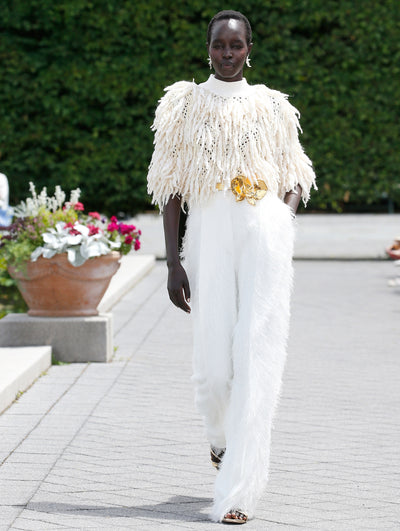 A model is wearing a monochromatic outfit consisting of a cream-colored, high-neck top with textured, fringe-like details and wide-leg, white trousers with a similar feathery texture.