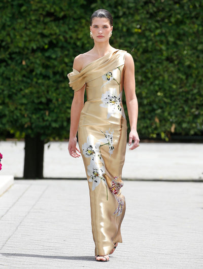 A model is wearing a gold, one-shoulder gown with a draped neckline and a fitted silhouette. The dress features large, elegant white floral prints with hints of green and purple.