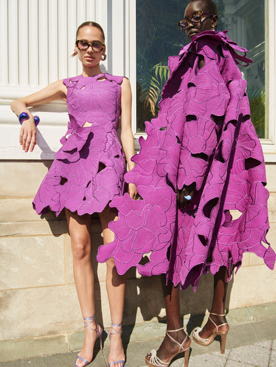 Two models pose outside in Oscar de la Renta ready-to-wear styles. One model is wearing a purple laser-cut floral dress and the other is wearing a purple laser-cut floral coat. 