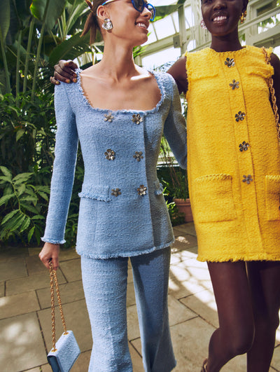 Two models pose in a green house in Oscar de la Renta ready-to-wear styles. One model is wearing a blue tweed set and the other is wearing a yellow tweed mini dress.