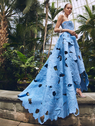A model poses in a green house on a ledge in an Oscar de la Renta laser cut floral blue gown. 