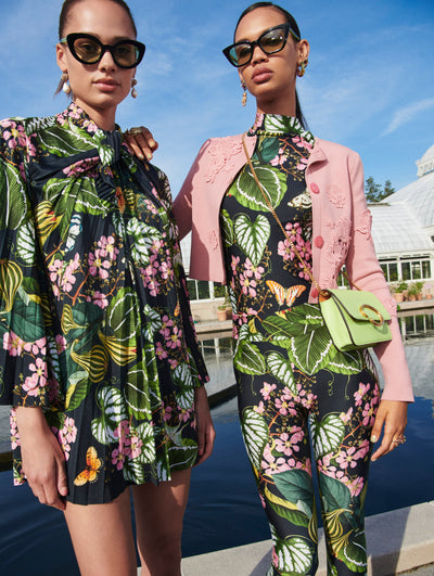 Two models pose outside in Oscar de la Renta ready-to-wear styles. One model is wearing a floral mini dress and the other is wearing a matching floral blouse and trouser set paired with a pink jacket. 