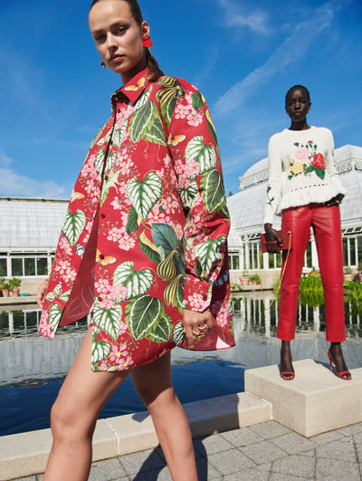Two models pose outside in Oscar de la Renta ready-to-wear styles. One model is wearing a red floral jacket and the other is wearing a floral pullover with red trousers. 