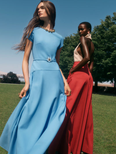 Two models are standing next to one another in the garden. The model on the left is wearing a powder blue midi cocktail dress with a floral belt. The model on the right is wearing a pink halter neck top and red flared pants. 