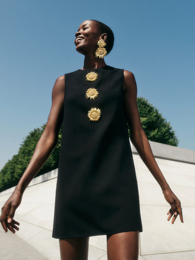 The model is wearing a classic shift dress, cut from black stretch-wool and finished with gold-tone sunflower brooches at the front.