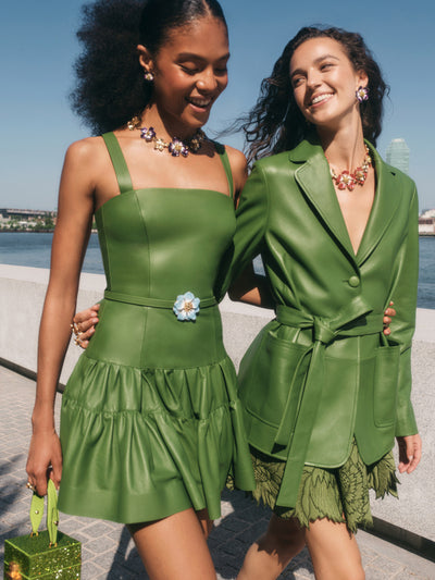 Two models are walking side by side. The model on the right is wearing a green leather jacket and green floral skirt with crystal floral necklace. The model on the left is wearing a green leather mini dress with a floral belt. 