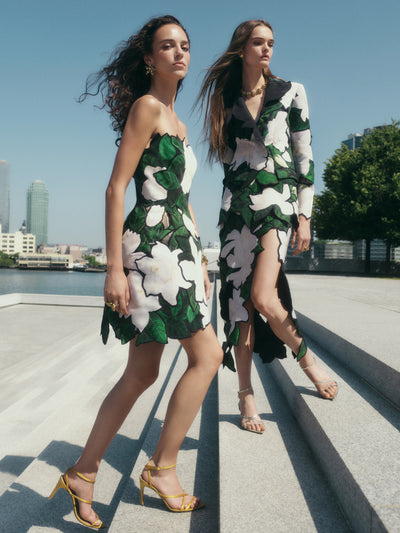 Two models are climbing stairs next to each other. The model on the left is wearing a strapless floral mini dress & the model on the right is wearing a matching floral skirt and jacket set. 