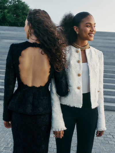 Two models stand next to each other against a flight of stairs. The model on the left is wearing a black and blue lace jacket & skirt set with an open back and the model on the right is wearing a white lace jacket and black pants. 