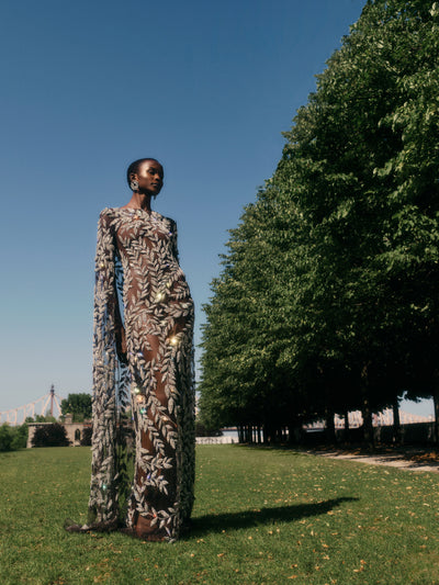 The model is wearing a tulle evening gown with crystal leaves. She poses in a garden with a row of trees to her right. 