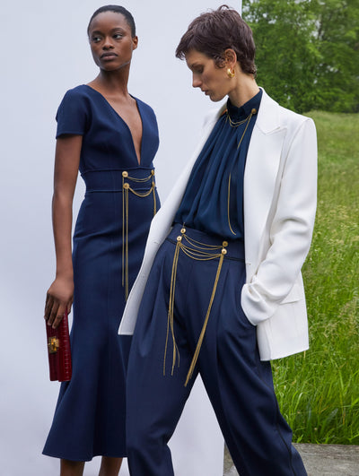 Two models; the model on the left wears a black dress with gold details and the model on the right wears a black top and pant with white suit jacket and gold details