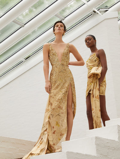 Two models; the model on the left wears a gold gown with flower print and the model on the right wears a short yellow dress with flower print