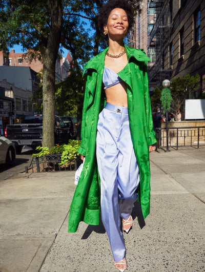 A model walks on the New York City sidewalk in an Oscar de la Renta green trench styled with a blue silk bralette top and trouser set. 