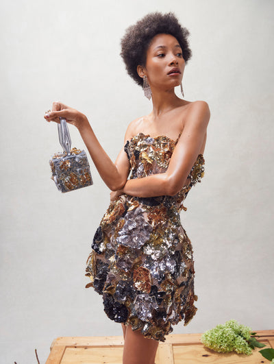 A model poses inside in an Oscar de la Renta gold and silver embroidered cocktail dress paired with a matching handbag. 