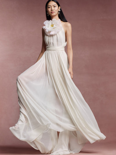 The model is wearing a halter neck white gown with an oversized flower at the neckline. 