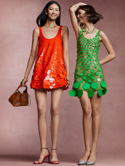 Two models stand next to each other against a pink backdrop. The model on the left is wearing an orange sequin mini dress and the model on the right is wearing a green crochet and sequin mini dress. 