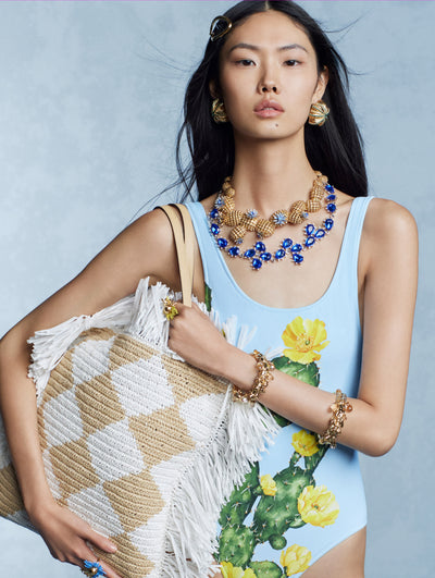 The model is wearing a blue one piece and multiple floral necklaces. 
