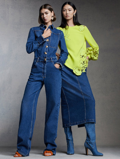 Two models are standing against a grey backdrop. The model on the left is wearing a denim jumpsuit with gold, floral buttons and the model on the right is wearing a yellow sweater with floral cutouts and a denim midi skirt. 