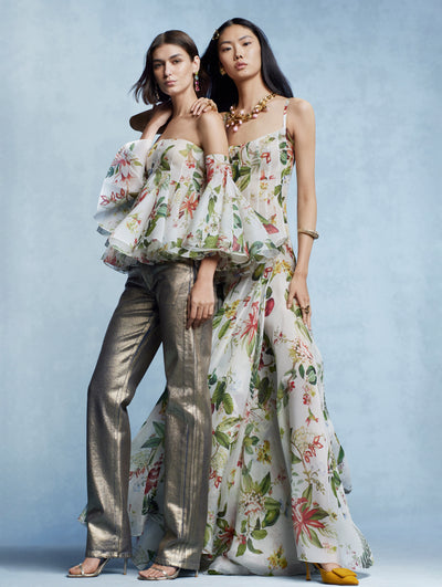 Two models stand together against a blue backdrop. The model on the left is in an off the shoulder top, adorned with the degrade Flora & Fauna motif and silver pants, and the model on the right is in a long degrade Flora & Fauna dress. 