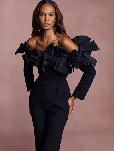 The model is wearing a black, off-shoulder blouse with bows at the neckline and black cigarette pants. 