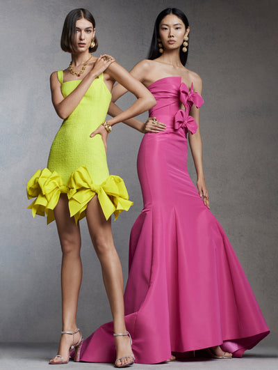 Two models stand together against a grey background. The model on the left is in a yellow mini dress with large bows on the hem and the model on the right is in a pink evening gown with bows at the bust. 