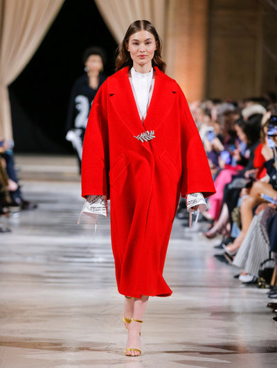 The model walks the runway in a red long-sleeve oversized wool coat with a crystal leaf pin closure, layered over a white long-sleeve blouse with silver sequin embroidered cuffs. Behind her, the blurred background shows the audience and another model in a black and white ensemble walking the runway.