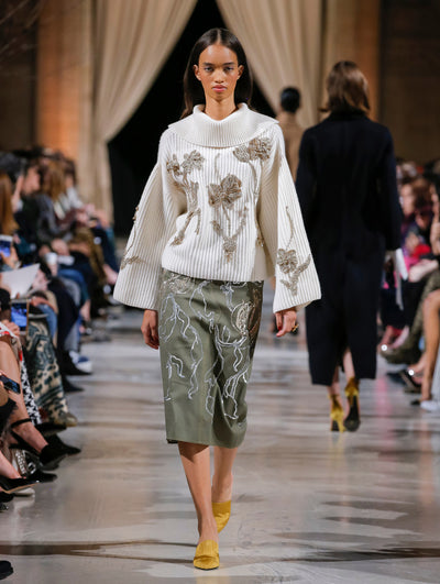 The model walks the runway in an ivory turtleneck pullover with three-dimensional crystal-embroidered florals and a khaki skirt with sequin-embroidered sinuous vines. Behind her, the blurred background shows the audience and two model walking the runway in the opposite direction.