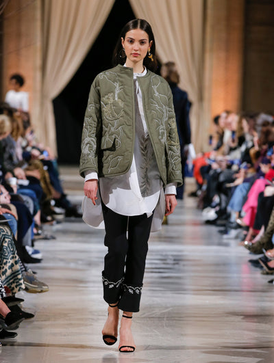 The model walks the runway in a khaki-hued bomber jacket with silver threadwork embroidered florals, layered over a white button-up long-sleeve blouse and black trousers with crystal-embroidered cuffs. Behind her, the blurred background shows the audience and another model in a navy ensemble walking the runway in the opposite direction.