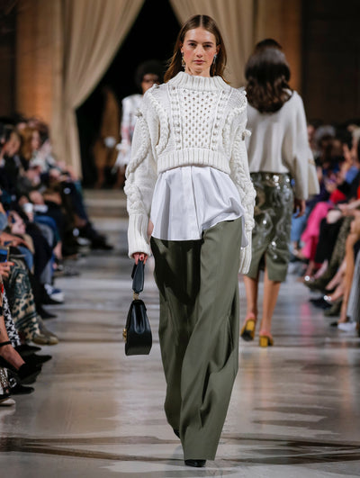 Holding a black top-handle handbag, the model walks the runway in a cream wide-ribbed cable knit pullover, layered over a white button-up blouse and khaki-hued trousers. Behind her, the blurred background shows the audience and several models walking the runway in various directions