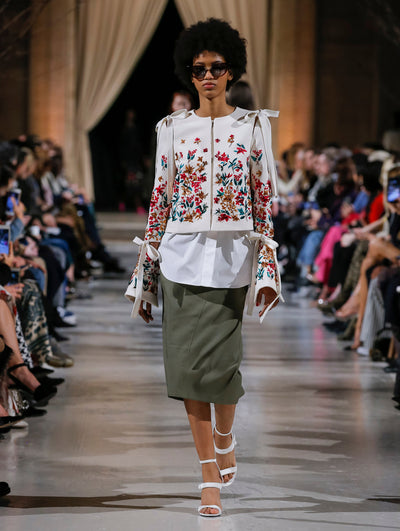 The model walks the runway in an ivory bow-detailed jacket with multi-colored sequin-embroidered florals, layered over a white button up blouse and khaki skirt. Behind her, the blurred background shows the audience and several models walking the runway in various directions.
