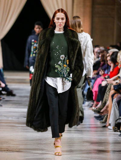 The model walks the runway in a green long-sleeve fur coat, layered over a green ribbed knit pullover with crystal-embroidered dandelions, a white button-up blouse with floral threadwork embroidery and navy trousers. Behind her, the blurred background shows the audience and two models walking the runway in various directions.