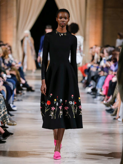 The model walks the runway in a black long-sleeve cocktail dress with multi-colored threadwork embroidered florals concentrated on the skirt. Behind her, the blurred background shows the audience and several models walking the runway in various directions. 