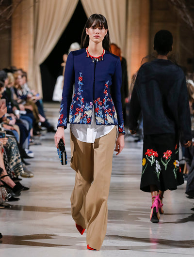 Holding a blue and black sequin-embroidered clutch, the model walks the runway in a navy long-sleeve jacket with multi-colored sequin embroidered florals, layered over a white button-up blouse and light brown wide-leg trousers. Behind her, the blurred background shows the audience and several models walking the runway in various directions. 