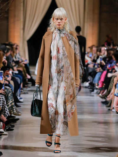 Holding a green tote bag, the model walks the runway in a light brown long-sleeve open-front wool coat, layered over a white silk chiffon tie-neck maxi dress printed with an all-over multi-colored floral design. Behind her, the blurred background shows the audience and two models walking the runway in various directions.