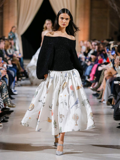 The model walks the runway in a black off-shoulder long-sleeve ribbed knit pullover and a white silk skirt printed with an all-over animal and floral design. Behind her, the blurred background shows the audience and another model in a white ensemble walking the runway.