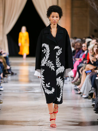 The model walks the runway in a black long-sleeve oversized wool coat with a crystal leaf pin closure and white leaf embroidery, layered over a white long-sleeve blouse with statement cuffs. Behind her, the blurred background shows the audience and another model in an orange ensemble walking the runway.