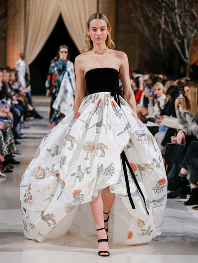 The model walks the runway in strapless high-low gown with a black bow-detailed bodice and white voluminous skirt printed with an all-over animal and floral design. Behind her, the blurred background shows the audience and two models in multi-colored ensembles walking the runway.