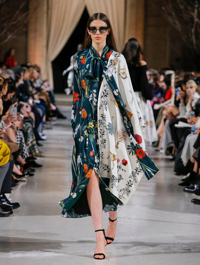 Wearing black sunglasses, the model walks the runway in a green and white color-block tie-neck silk caftan printed with an all-over floral design. Behind her, the blurred background shows the audience and several models walking the runway in various directions.