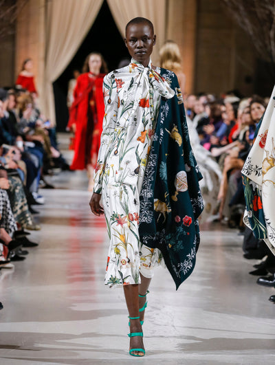 The model walks the runway in a green and white color-block tie-neck silk dress printed with an all-over floral design. Behind her, the blurred background shows the audience and several models walking the runway in various directions.