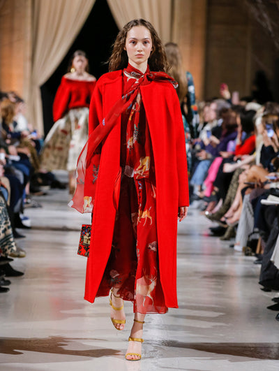 The model walks the runway in a red long-sleeve open-front oversized wool coat, layered over a red tie-neck silk dress printed with an all-over animal design. Behind her, the blurred background shows the audience and several models walking the runway in various directions.