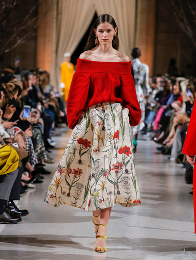 The model walks the runway in a red off-shoulder long-sleeve ribbed knit pullover and a beige wool skirt printed with an all-over floral design. Behind her, the blurred background shows the audience and several models walking the runway in various directions.