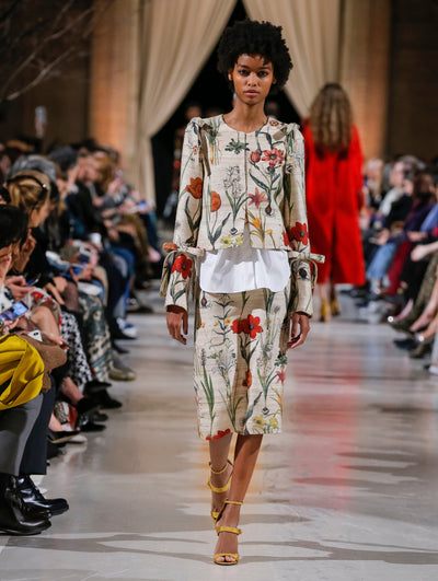 The model walks the runway in a beige bow-detailed jacket printed with an all-over floral design, layered over a white button-up blouse and coordinating beige wool skirt printed with an all-over floral design. Behind her, the blurred background shows the audience and several models walking the runway in various directions.