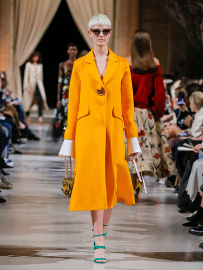 Holding a gold gardenia appliqué handbag and wearing rouge sunglasses, the model walks the runway in an orange long-sleeve wool coat with a crystal bird pin closure, layered over a white long-sleeve blouse. Behind her, the blurred background shows the audience and several models walking the runway in various directions.