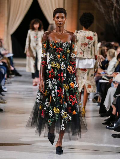 The model walks the runway in a black illusion-neck long-sleeve tulle evening gown with multi-colored sequin-embroidered florals. Behind her, the blurred background shows the audience and several models walking the runway in various directions.