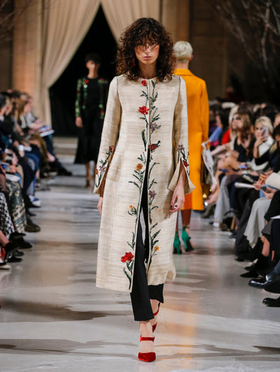 The model walks the runway in a beige long-sleeve wool coat with multi-colored crystal-embroidered florals concentrated on the split sleeves and front. Behind her, the blurred background shows the audience and several models walking the runway in various directions.