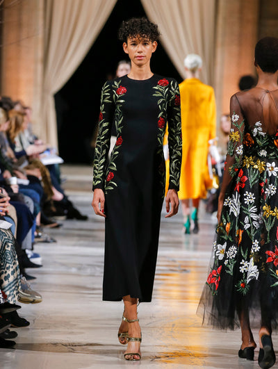 The model walks the runway in a black long-sleeve pencil cocktail dress with multi-colored sequin-embroidered florals concentrated on the sleeves and sides. Behind her, the blurred background shows the audience and several models walking the runway in various directions.